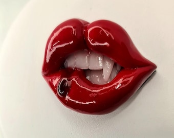 May include: A pair of red, glossy lips with a silver ring piercing the bottom lip. The lips are slightly parted, revealing a set of white teeth.