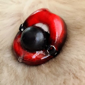 Pode incluir: Uma boca esculpida vermelha e brilhante com uma mordaça de bola preta. A boca está aberta e mostra dentes brancos. A mordaça está presa a uma correia preta.