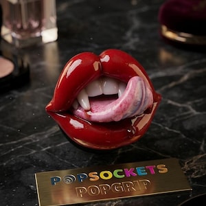 Könnte beinhalten: Ein glänzender, lippenförmiger PopSockets PopGrip mit weißen Vampirzähnen und einer rosa Zunge. Darunter befindet sich eine rechteckige, goldfarbene Metallplatte mit der eingravierten Aufschrift POPSOCKETS POPGRIP in bunten und goldenen Buchstaben.