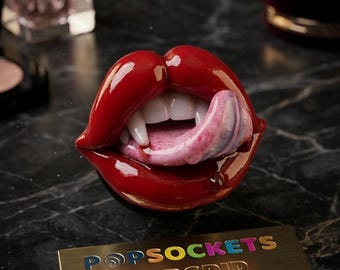 May include: A glossy red lip-shaped PopSockets PopGrip featuring white vampire fangs and a pink tongue. Below the accessory is a rectangular gold-toned metal plate with the text POPSOCKETS POPGRIP engraved in multi-colored and gold lettering.