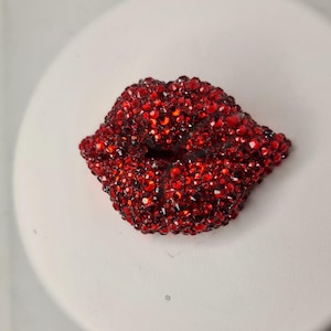 May include: A decorative accessory in the shape of red lips, covered in sparkling jewels. The lips are a deep red hue, with each jewel reflecting light. This item could be used as an embellishment.