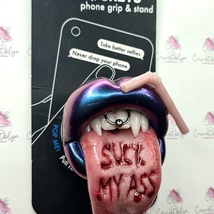 Puede incluir: Un soporte y soporte para teléfono PopSockets con una boca estilizada con colmillos, un labio perforado y una lengua con las palabras "SUCK MY ASS" en rojo. La boca es una mezcla de azul y morado.