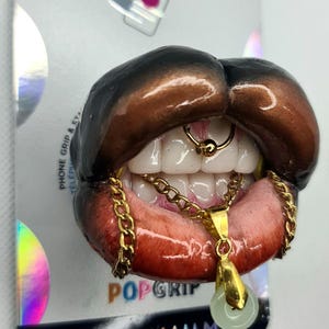 Puede incluir: Un soporte para tel&eacute;fono con una boca abierta esculpida, labios marr&oacute;n oscuro, dientes blancos y un aro de septum dorado. Una cadena dorada cuelga de los dientes, sosteniendo un peque&ntilde;o colgante circular de color verde p&aacute;lido sobre una tarjeta.