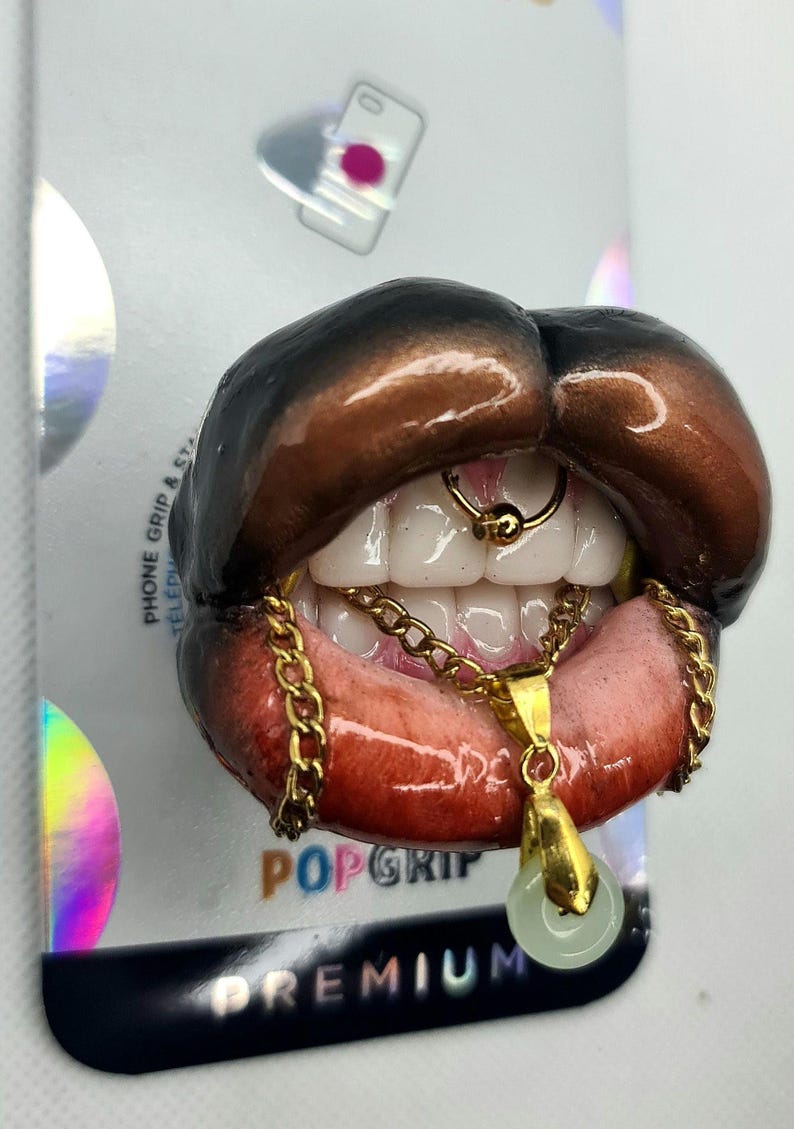 Puede incluir: Un soporte para tel&eacute;fono con una boca abierta esculpida, labios marr&oacute;n oscuro, dientes blancos y un aro de septum dorado. Una cadena dorada cuelga de los dientes, sosteniendo un peque&ntilde;o colgante circular de color verde p&aacute;lido sobre una tarjeta.