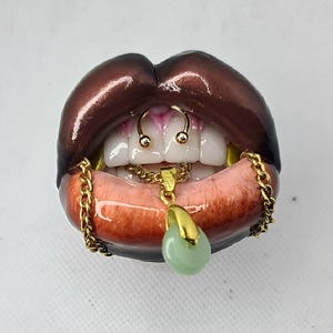 Puede incluir: Una escultura de labios de color marr&oacute;n oscuro con dientes blancos, que presenta un anillo de septum dorado, una cadena dorada sobre el labio inferior y un colgante dorado con una piedra lisa de color verde p&aacute;lido en el centro.