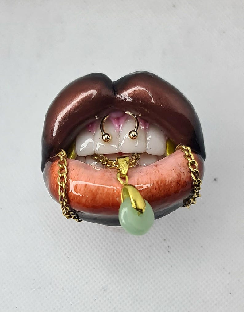 Puede incluir: Una escultura de labios de color marr&oacute;n oscuro con dientes blancos, que presenta un anillo de septum dorado, una cadena dorada sobre el labio inferior y un colgante dorado con una piedra lisa de color verde p&aacute;lido en el centro.