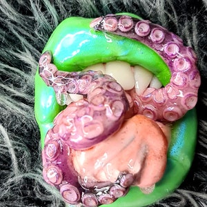May include: A green and purple octopus tentacle with suckers is wrapped around a pink and white tongue. The tongue is sticking out of a green mouth with white teeth.
