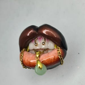 Puede incluir: Una escultura de labios de color marr&oacute;n oscuro con dientes blancos y un anillo de septum dorado. Una cadena dorada cuelga de la boca, sosteniendo un peque&ntilde;o colgante redondo de color verde claro. Los labios tienen un acabado brillante.