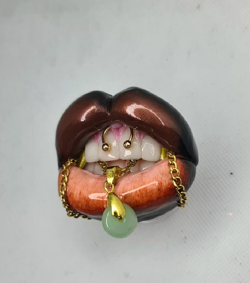 Puede incluir: Una escultura de labios de color marr&oacute;n oscuro con dientes blancos y un anillo de septum dorado. Una cadena dorada cuelga de la boca, sosteniendo un peque&ntilde;o colgante redondo de color verde claro. Los labios tienen un acabado brillante.