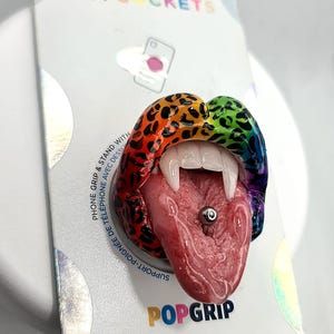 Puede incluir: Un PopGrip PopSockets con un diseño de boca abierta y colorida, con colmillos y un piercing en la lengua. El agarre presenta un estampado de leopardo arcoíris y está fijado a un respaldo blanco con los logotipos de PopSockets y PopGrip.