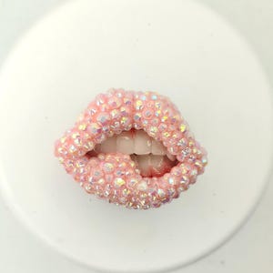 Puede incluir: Un objeto decorativo en forma de labios rosa, adornado con joyas. Los labios están cubiertos de pedrería iridiscente, con dientes visibles y una boca ligeramente abierta. El objeto está sobre un fondo blanco.