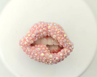 May include: A pink, bedazzled lips-shaped decorative item. The lips are covered in iridescent rhinestones, with visible teeth and a slight open mouth. The item is set against a white background.