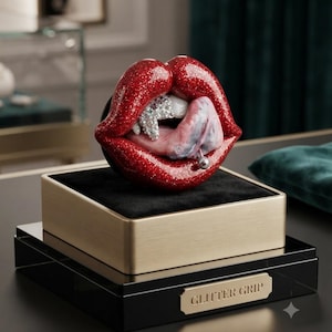 May include: A red glittery lips sculpture with a tongue and silver-toned teeth accents, mounted on a black and gold square base. The base features a gold-toned plaque with the text GLITTER GRIP engraved in black capital letters.