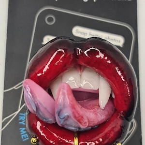 Puede incluir: Un soporte y agarre para teléfono PopSockets con un diseño de boca roja brillante, con colmillos y una lengua rosa. Tiene un aro labial dorado y el texto "TRY ME! Pull twice to expand".