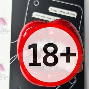 Puede incluir: Agarre y soporte para teléfono rojo con acabado brillante en forma de labios. El soporte tiene un círculo blanco con el texto "18+" en negro.