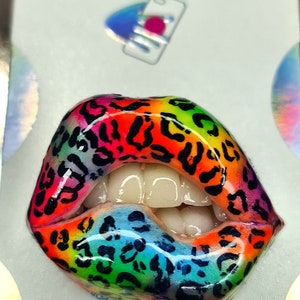 Puede incluir: Una agarradera de teléfono colorida con un diseño de estampado de leopardo arcoíris. La agarradera tiene forma de labios con dientes blancos. El texto "POPGRIP" es visible en la parte inferior de la imagen.