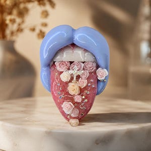 Puede incluir: Un adorno escultórico con labios azul bígaro brillantes, dientes blancos y una lengua rosa texturizada. La lengua está decorada con pequeñas rosas de color rosa pálido, una cruz plateada central y pedrería transparente dispersa.