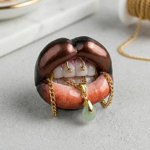 Puede incluir: Una escultura de labios abiertos con pintalabios marr&oacute;n oscuro, dientes blancos y un piercing de septum dorado. Una cadena dorada cuelga de los dientes, sosteniendo un peque&ntilde;o colgante en forma de l&aacute;grima de color verde p&aacute;lido. Acabado brillante.