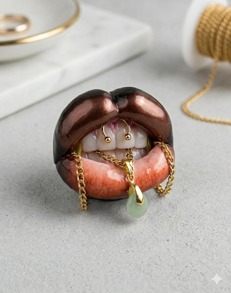 Puede incluir: Una escultura de labios abiertos con pintalabios marr&oacute;n oscuro, dientes blancos y un piercing de septum dorado. Una cadena dorada cuelga de los dientes, sosteniendo un peque&ntilde;o colgante en forma de l&aacute;grima de color verde p&aacute;lido. Acabado brillante.