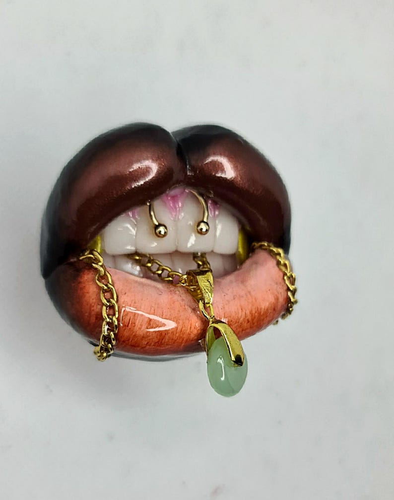 Puede incluir: Una escultura de labios abiertos con bordes exteriores de color marr&oacute;n oscuro y un &aacute;rea interior de color rosa rojizo. Los dientes blancos presentan un piercing de septum dorado y una cadena dorada sobre los dientes con un colgante ovalado verde.