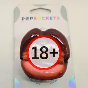 Puede incluir: Un accesorio para teléfono PopGrip de color rojo y rosa con un diseño 3D de un par de labios. El embalaje tiene el texto "PopSockets" y "PopGrip" y una advertencia de 18+.