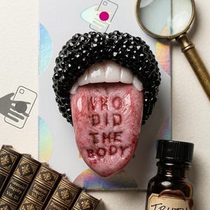 May include: A PopSockets grip with a tongue design, the words "WHO DID THE BODY" are engraved. The lips are encrusted with black gems. A small bottle labelled "TRUTH" is also visible.