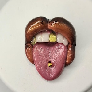 Puede incluir: Escultura detallada de una boca con labios marrones, dientes blancos y grillz dorados. La lengua rosa tiene un piercing de barra dorado. La escultura está sobre una superficie blanca.