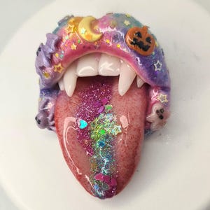 May include: A sculpted mouth ornament featuring glossy lips in a gradient of pink, purple, and blue with embedded glitter, stars, a crescent moon, a pumpkin, and small ghosts. The mouth has white fangs and a tongue covered in pink and iridescent glitter.