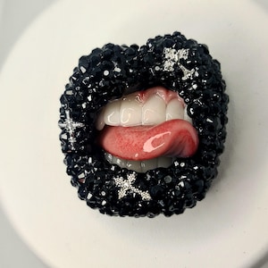 Puede incluir: Un diseño decorativo de labios abiertos, adornado con elementos negros en forma de cristal. La lengua está extendida y los dientes son visibles. Acentos plateados en forma de cruz están colocados en los labios.
