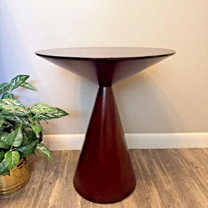 May include: A dark brown side table featuring a circular top and a conical base. The table has a smooth, solid finish and a flared silhouette where the wide, flat top meets the narrow neck of the base, which then widens toward the floor.