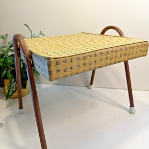 May include: A vintage footstool with a yellow patterned top and brown legs. The rectangular top features a repeating pattern in black and brown. The legs are brown with white capped feet and curved brown handles.
