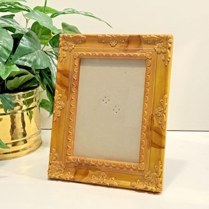 May include: A rectangular picture frame with a light brown wood grain finish and ornate, raised floral carvings along the edges. The frame features a scalloped inner border surrounding a plain, light beige insert with two small, centered cutouts.