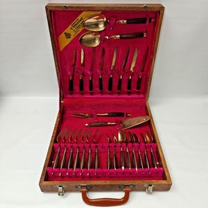May include: A wooden storage case with a bright pink velvet interior containing a set of gold-toned flatware with dark brown handles. The set includes various spoons, forks, and knives, with a label reading V. THAILAND in the upper corner.