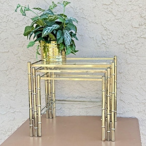 May include: A set of three gold-coloured nesting tables with a bamboo-style design. Each table has a clear glass top. A gold-coloured planter with a green plant sits on the largest table. The tables are arranged in a nested configuration.
