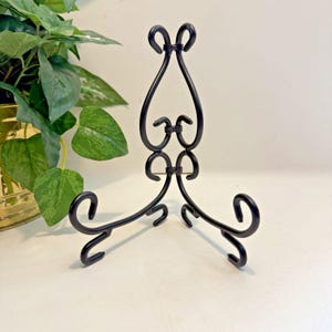 May include: A black metal display stand featuring a curved, scrollwork design with two base supports and a central upright frame. The stand is constructed from thin, rounded metal rods that form symmetrical loops and decorative swirls throughout.
