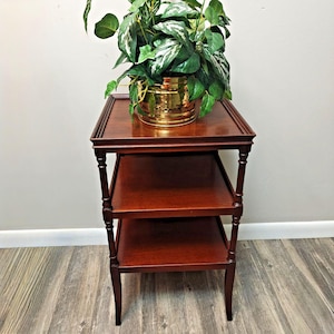 May include: A three-tiered wooden side table with a dark brown finish and four slender legs. The top surface holds a shiny gold-toned metal planter containing a lush green plant with variegated leaves. The table features a square design.