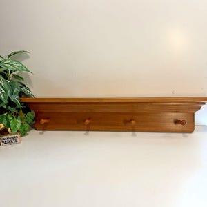 May include: A rectangular wooden wall shelf featuring a flat top surface and four evenly spaced wooden pegs extending from the front panel. The shelf is finished in a medium brown wood tone with a smooth, polished texture and a simple moulding design.