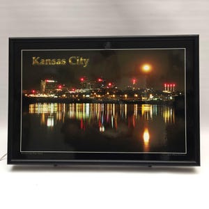 May include: A framed photograph of the Kansas City skyline at night, featuring illuminated buildings and their reflections on the water. The text Kansas City is printed in gold in the upper left corner, enclosed in a simple black frame.