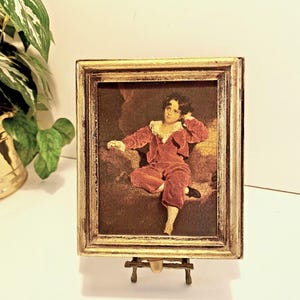 Op de afbeelding: Een ingelijste prent van een jong persoon in een rood fluwelen pak, op een kleine gouden ezel. De prent heeft een gouden lijst met een distressed afwerking. Het kunstwerk toont een zittende persoon.