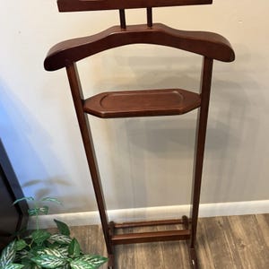 May include: A dark brown wooden valet stand featuring a wide contoured hanger at the top for garments, a horizontal bar above it, and a central rectangular tray with rounded edges for small items, supported by two vertical legs and a base.