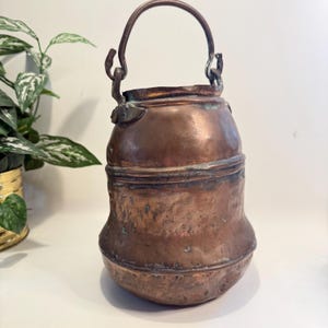 May include: A vintage copper bucket with a curved handle. The bucket has a rounded base and a cylindrical body with a slightly flared top. The copper has a rich, aged patina, with areas of darker oxidation. The handle is attached with decorative brackets.
