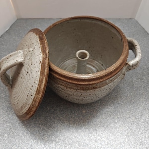 May include: A speckled beige ceramic garlic roaster with a removable lid. The round pot features two side handles, a brown rim, and a central vertical spike inside. The lid has a matching handle and brown rim, showing a textured clay finish.
