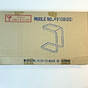 May include: A rectangular cardboard shipping box featuring a blue line drawing of a C-shaped side table. The text reads FRAGILE HANDLE WITH CARE, MODLE NO. #910B(GS), and MODEL NO. #910B(GS) MADE IN TAIWAN. The box is tan with dark blue text.