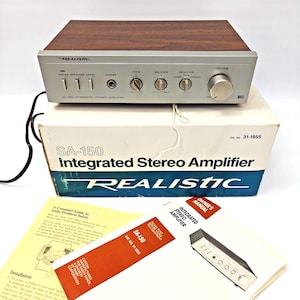 Puede incluir: Un amplificador estéreo integrado Realistic SA-150 con panel frontal plateado, parte superior de madera y perillas de control. Incluye la caja de cartón blanca original marcada como Cat. No. 31-1955, un manual de usuario y una guía de seguridad.