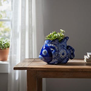 May include: A glossy ceramic fish-shaped planter featuring a deep blue and white intricate floral pattern. The fish has a rounded body, a prominent eye, and a wide tail fin. A small green succulent plant is nestled inside the top opening.
