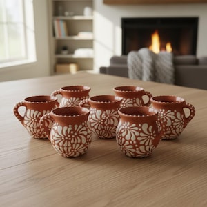 Puede incluir: Un juego de seis tazas de color terracota con forma redondeada y un patrón floral blanco pintado en el exterior. Cada taza tiene un asa y un borde festoneado con pequeños puntos blancos, dispuestas en grupo sobre una superficie de madera.