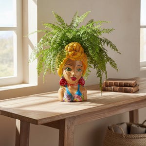 May include: A ceramic head-shaped planter featuring a person with a yellow turban, green eyes, and red lips. The figure wears a blue and pink top with large red earrings. A lush green fern plant grows out of the top of the head, serving as hair.
