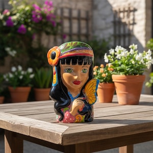 May include: A ceramic head-shaped planter featuring a person with long black hair, green eyes, and red lips. The figure wears a colorful headband with an orange flower and holds a yellow butterfly near their face. The base has pink and floral patterns.
