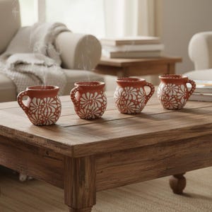May include: Four small, round terracotta mugs with a handle, each featuring a hand-painted white floral pattern. The mugs are arranged in a row on a wooden surface, with wisps of steam rising from the first mug on the left, indicating warmth.