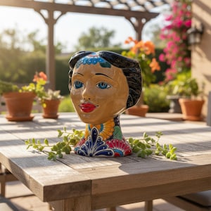 May include: A ceramic head-shaped planter featuring a glossy tan complexion, black hair, blue eyes, and red lips. The base is decorated with vibrant, multi-colored floral and geometric patterns in shades of blue, yellow, red, and green.